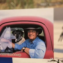 Man wearing a hat and glasses sitting in the driver's seat of a red vehicle with a dog beside him